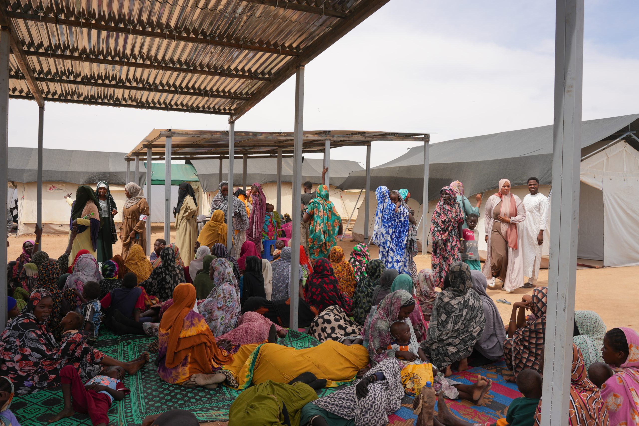 Patients waiting in the triage area, seated in the shade. The clinic in Tine transit camp provides essential healthcare services, vaccinations, clean dressings for trauma wounds, malnutrition care, and care for pregnant women and victims and survivors of sexual violence. ©️ Julie David de Lossy/MSF